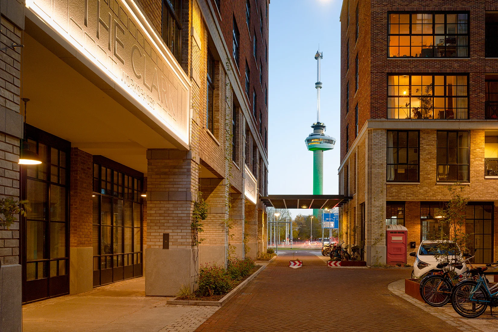 Doorkijk vanaf de Karl Weisbardstraat in Rotterdam met uitzicht op de verlichte Euromast tussen moderne bakstenen gebouwen.